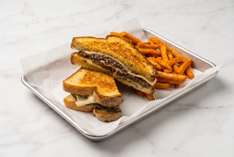 Grilled cheese sandwich with ground beef and fries on a metal tray with white paper.